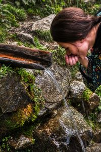 Woman drinking water flowing from stream by rock in forest