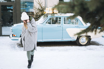 Young woman in warm clothes with cup of coffee in hands near retro car decorated christmas tree 