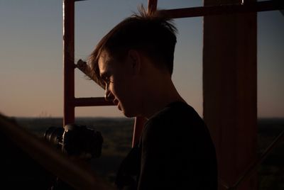 Side view of teenage boy holding camera amidst metallic structure