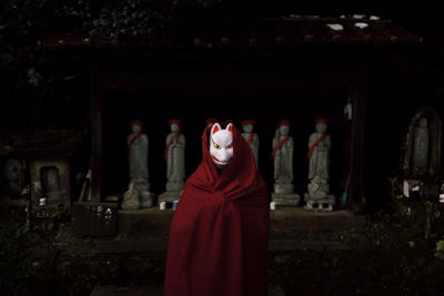 Person in fox mask standing in front of statues at night