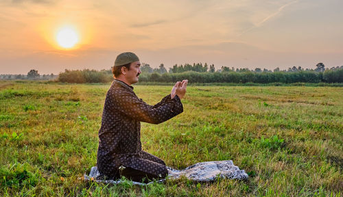 A muslim senior man wearing a skullcap and traditional clothes prays at sunset in a field