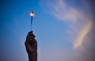 Cropped hand holding sparkler burning against blue sky during sunset