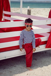 Stylish baby boy in a striped t-shirt and sunglasses stands at the red beach sunbed