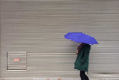 Side view of woman holding purple umbrella while walking by wall