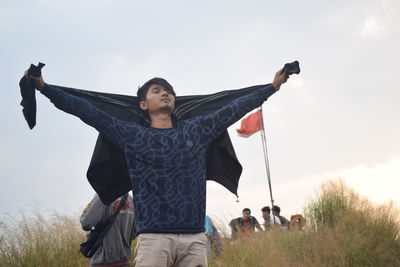 Young man with arms outstretched while standing on field against sky