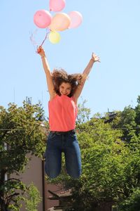 Happy young woman with balloon jumping against sky during sunny day