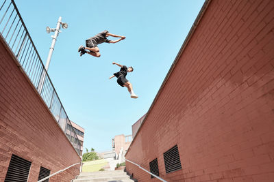 From below side view of strong males doing parkour and jumping from metal railing on brick building while showing stunts in city