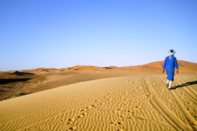 Man walking on sand dune in desert against clear blue sky