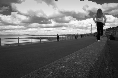 Rear view of girl walking on retaining wall against cloudy sky