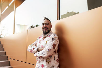 Bearded man leaning on wall with arms crossed against office building