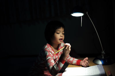 Cute girl studying at table with illuminated lamp in darkroom