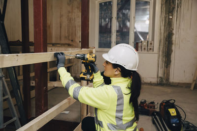 Female worker in hardhat using drill on wooden plank while working at construction site