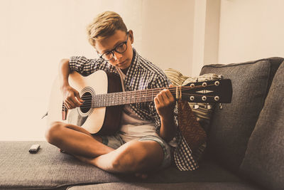 Cute boy playing guitar while sitting on sofa at home