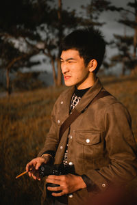 Young man holding camera while standing on land against trees