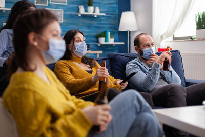 Cheerful people wearing mask drinking beer while watching tv at home