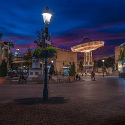 Illuminated street by buildings against sky at night