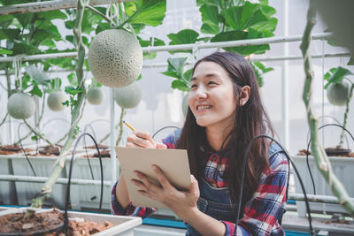 Beautiful smiling woman writing in book while looking at melon at farm