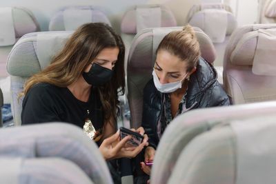 Woman with mask showing smartphone to friend in airplane