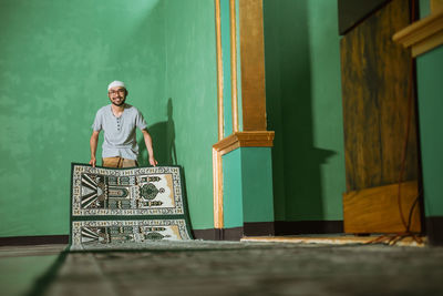Happy young man with carpet standing against wall in mosque