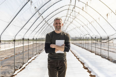 Smiling farmer with arms crossed holding digital tablet in agricultural field