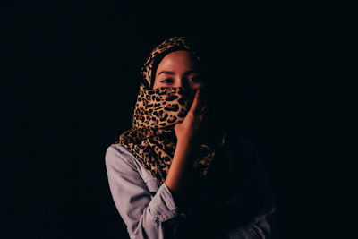 Portrait of young woman covering face with headscarf against black background