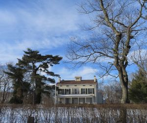 Bare trees on field by house against sky