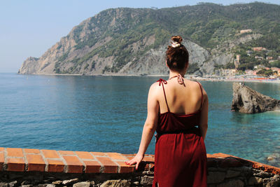 Rear view of woman wearing red dress while standing by retaining wall against sea