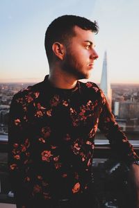Young man looking away in city against sky during sunset