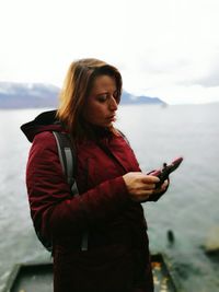 Serious woman looking at mobile phone on monopod while standing against lake during winter