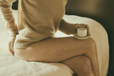 Woman in a camel sweater having a coffee on the side of the bed ii