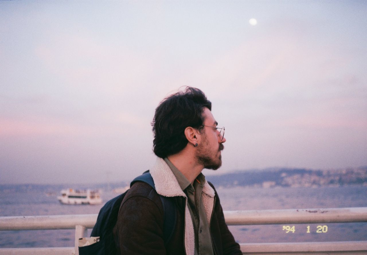 SIDE VIEW OF YOUNG MAN LOOKING AWAY AGAINST SKY