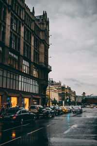 Cars on road by buildings in city against cloudy sky