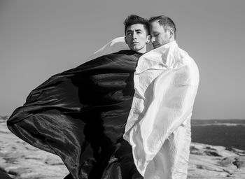 Young gay couple covered in textile standing on beach against sky