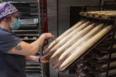 Side view of focused female baker in protective mask making notches on raw baguettes placed on baking pan in bakehouse