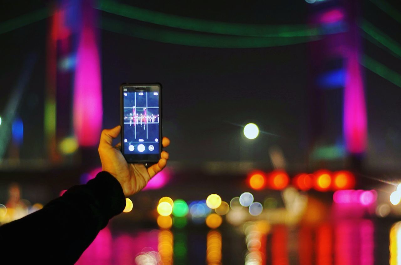 human hand, illuminated, night, human body part, real people, photographing, holding, focus on foreground, multi colored, wireless technology, technology, communication, one person, photography themes, mobile phone, built structure, architecture, close-up, lifestyles, building exterior, outdoors, sky, people