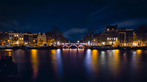 Illuminated bridge over river by buildings against sky at night