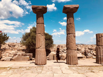 Full length of woman with arms raised standing by old ruins against blue sky