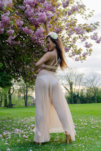 Rear view of young woman in sheer clothing posing by cherry blossoms at park
