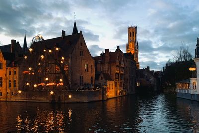 Illuminated buildings by river against cloudy sky