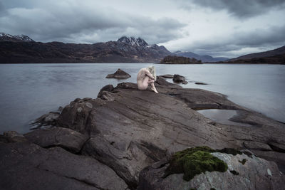 Naked woman on rock by lake against sky
