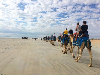 People riding on camels at beach against cloudy sky