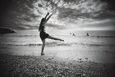 Full length of woman with arms raised standing on one leg at beach against cloudy sky