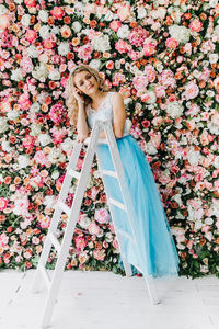 Young woman wearing blue dress standing on ladder against flowering plants