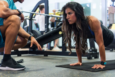 Focused ethnic female athlete doing push ups on mat under control of crop unrecognizable personal trainer in modern gym