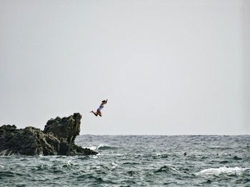 Teenage girl jumping from rock into sea against sky