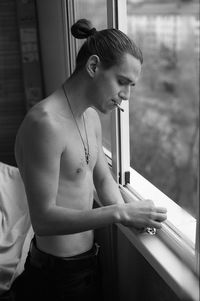 Shirtless young man smoking cigarette by window at home