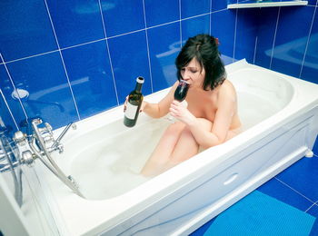 High angle view of naked woman drinking wine while sitting in bathtub