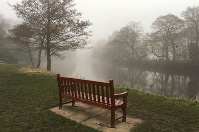 Empty bench by calm lake in foggy weather
