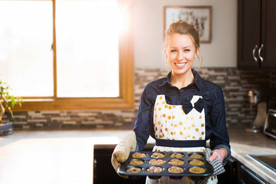 Portrait of smiling young woman holding baking sheet in kitchen at home