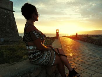 Young woman sitting on retaining wall by footpath during sunset
